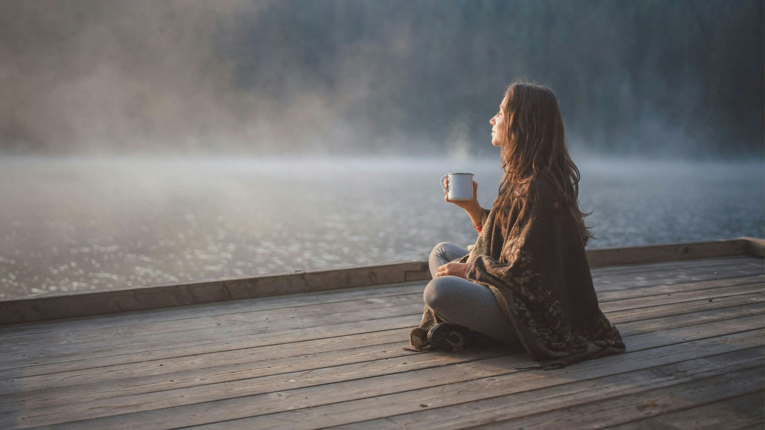 Femme sereine, assise en tailleur sur un ponton, qui contemple le paysage au petit matin et déguste un bon thé chaud.